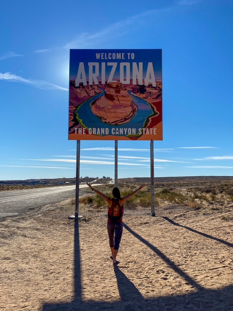 Sara standing in front of the Welcome to Arizona The Grand Canyon State sign with her hands raised high