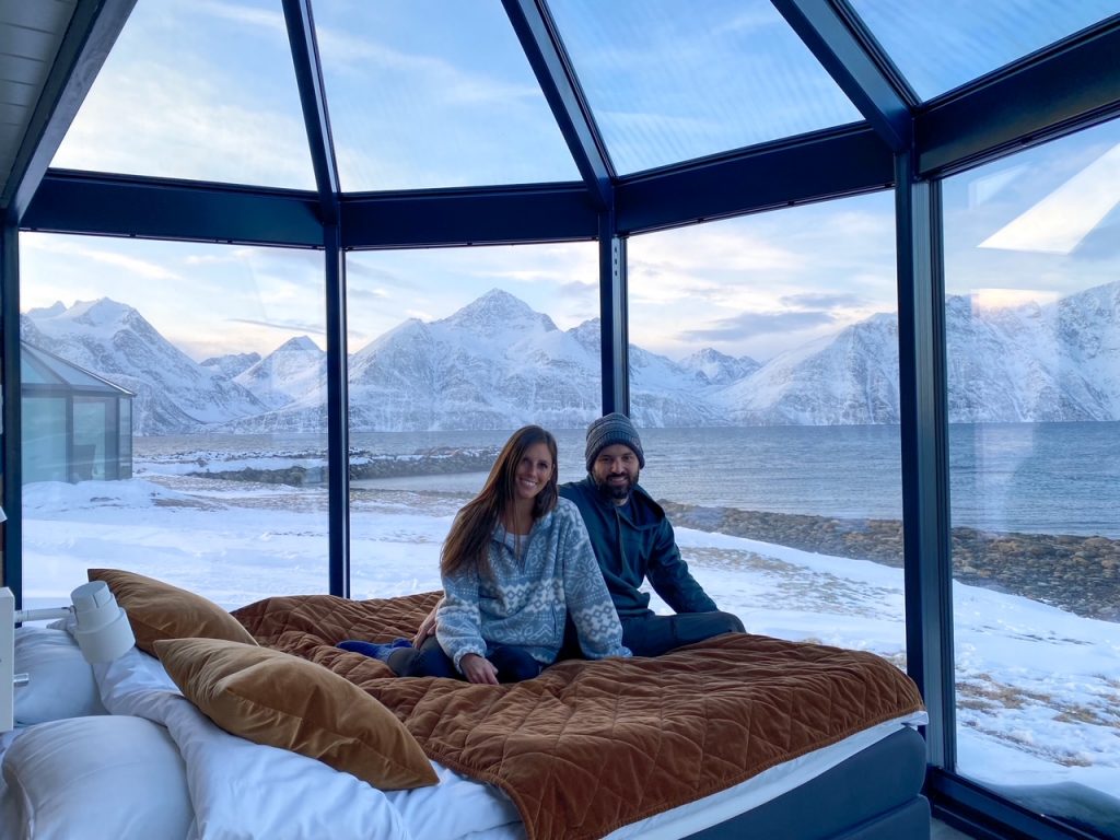 Sara and Tim sitting on the bed in their glass igloo overlooking the Lyngen Alps during the winter at Lyngen North in Rotsund, Norway