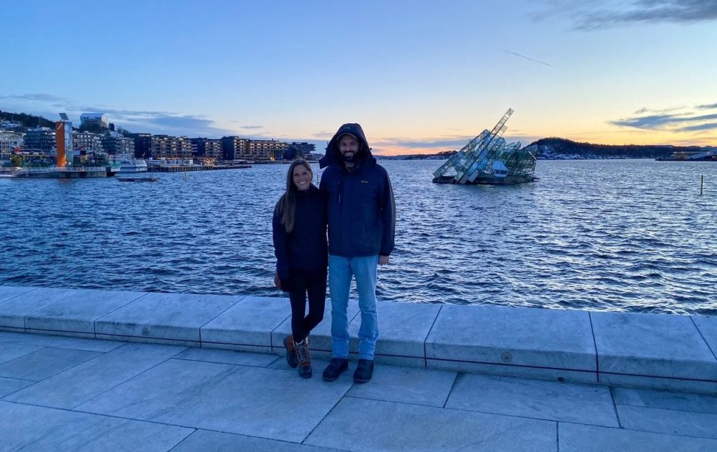 couple standing in front of the Oslo Fjord at sunset with the sailboat/iceberg-shaped “She Lies” sculpture in the background